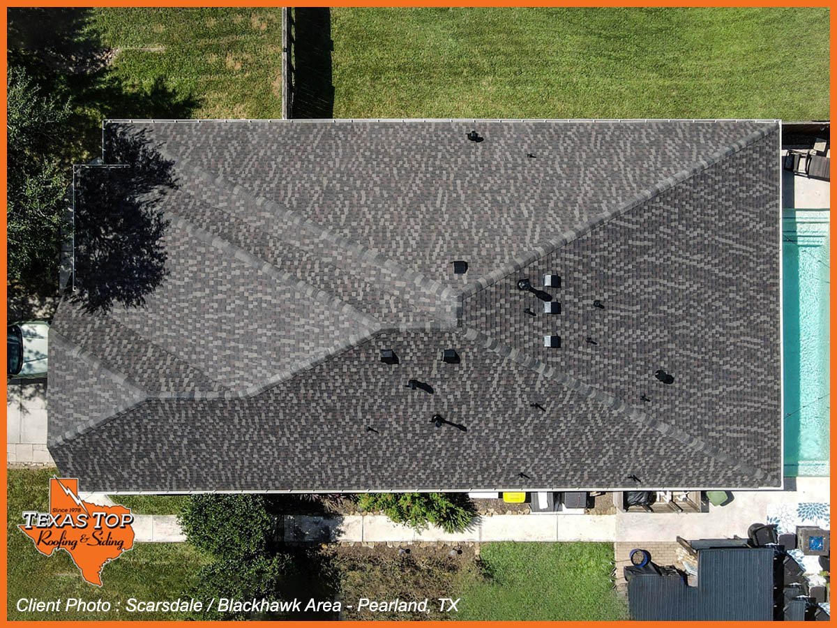 Aerial view of a residential driveway with dark asphalt shingles and scattered debris, surrounded by green lawn and trees