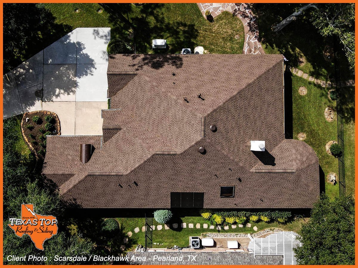 Aerial view of a brown brick residential home with complex rooflines, surrounded by green landscaping and driveway