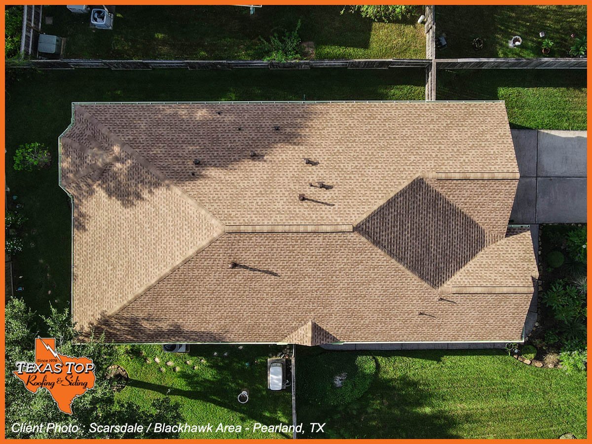 Aerial view of a residential house with a tan and gray shingled roof surrounded by green lawn and driveway