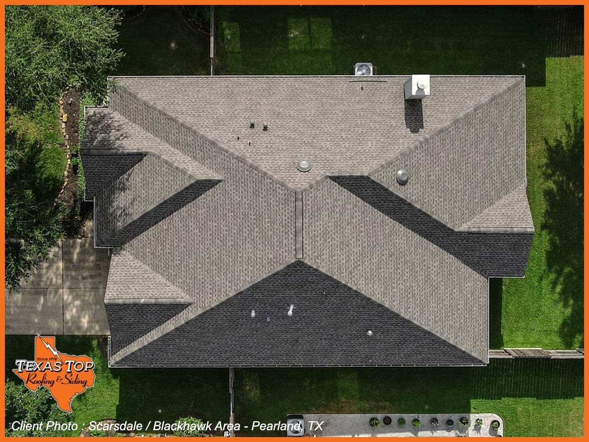 Aerial view of a residential house with gray and black roof, surrounded by green lawn and trees